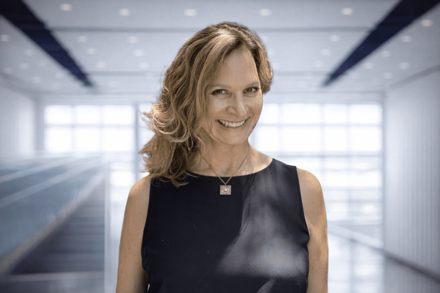 HeatherBeck-DirectorofBusinessDevelopment Woman with wavy hair wearing a sleeveless black top and a necklace, smiling, standing in a bright modern indoor setting with large windows.