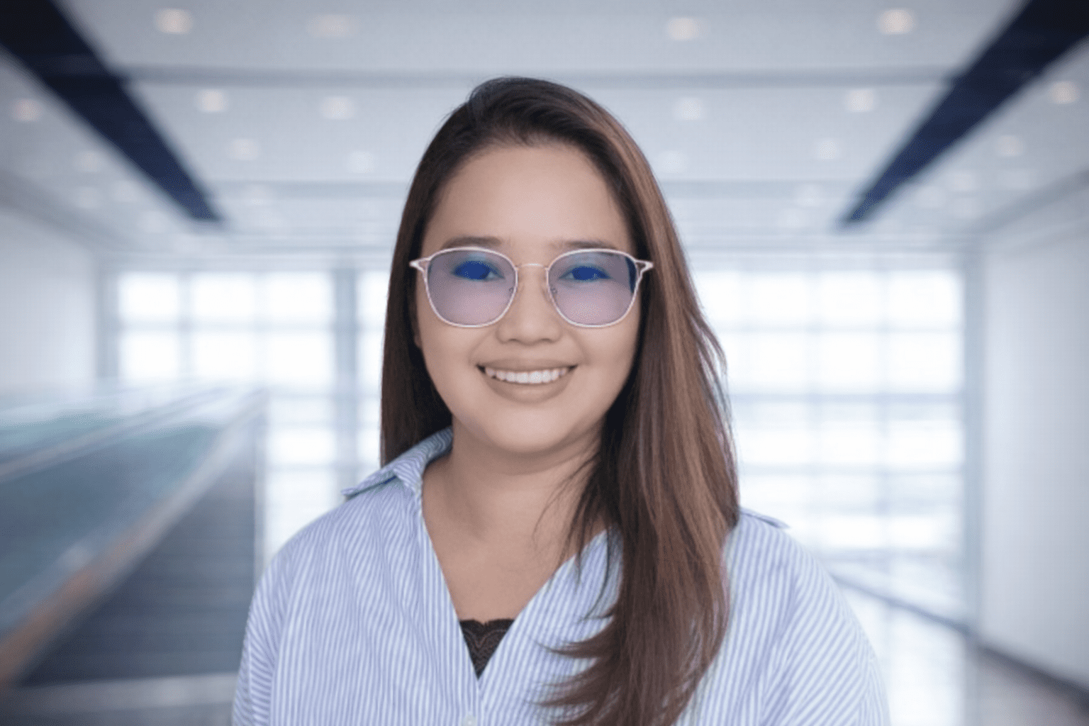 MarizSoliva-HeadofAdministration A woman with long brown hair and blue glasses smiles at the camera, standing in a modern, bright indoor setting with large windows.