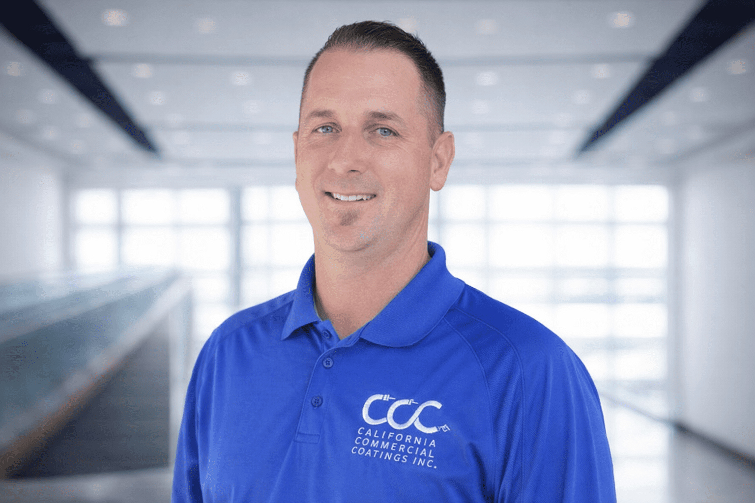 MattSwall-DirectorofProduction A man in a blue "California Commercial Coatings Inc." polo shirt stands indoors, smiling at the camera with a blurred modern background.
