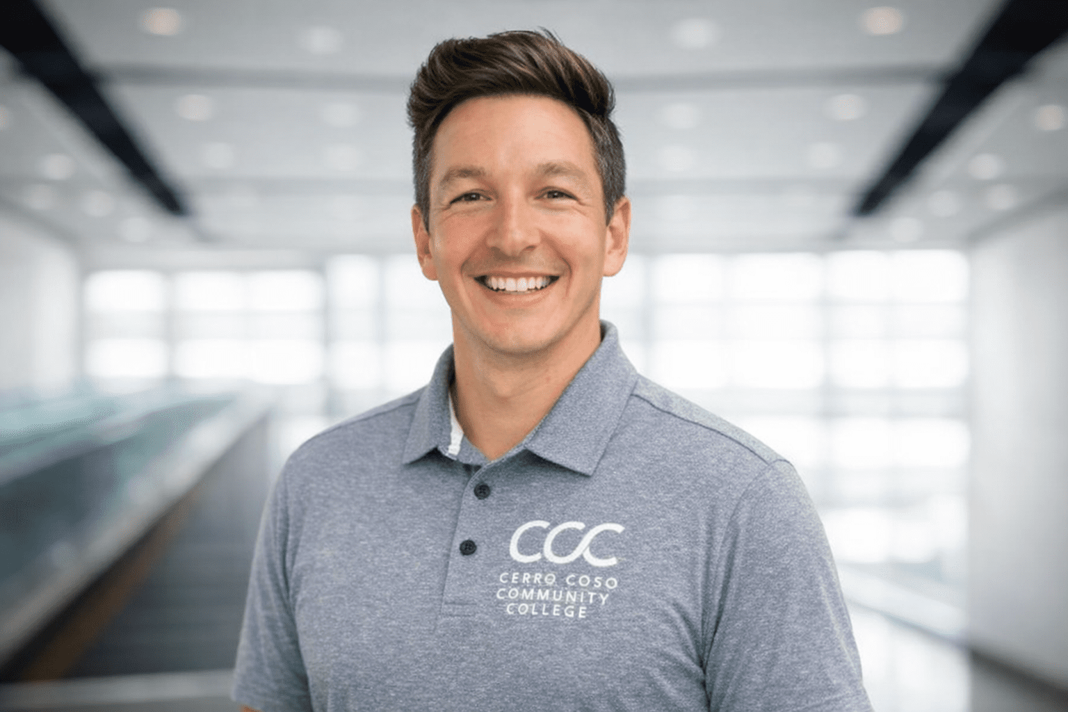 TyHaga-President A smiling person wearing a light blue Cerro Coso Community College polo shirt stands indoors with a blurred, bright background.