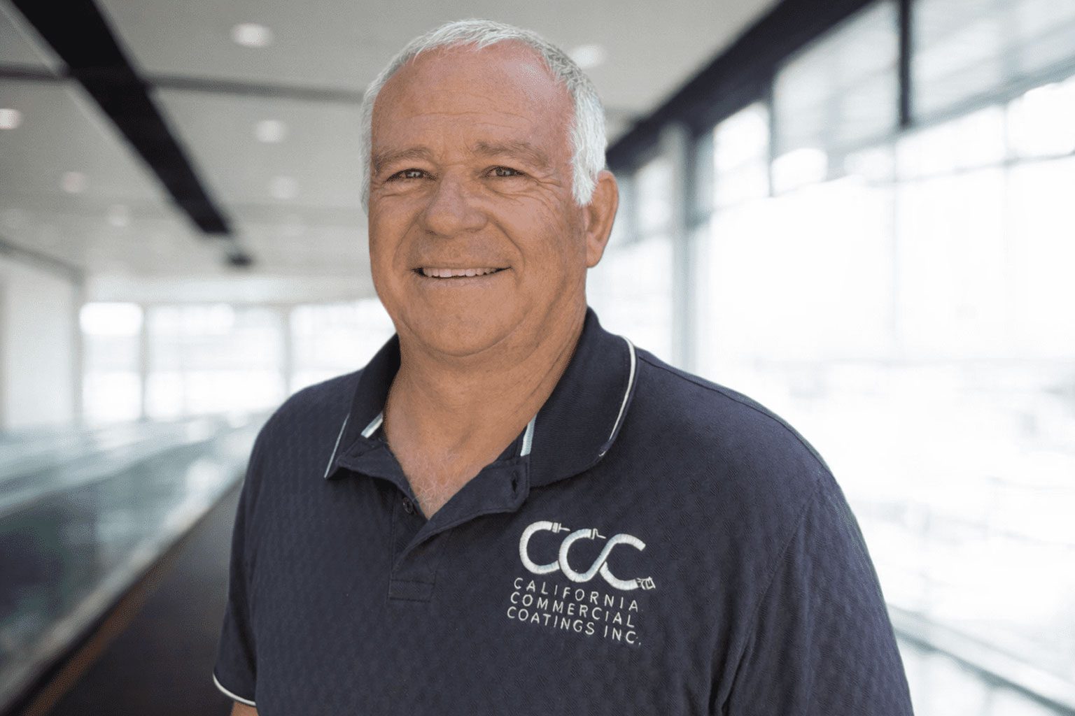 MarkTretter-DirectorofWaterproofing-WEB A man with short gray hair wearing a navy blue "California Commercial Coatings Inc." polo shirt stands indoors near large windows.